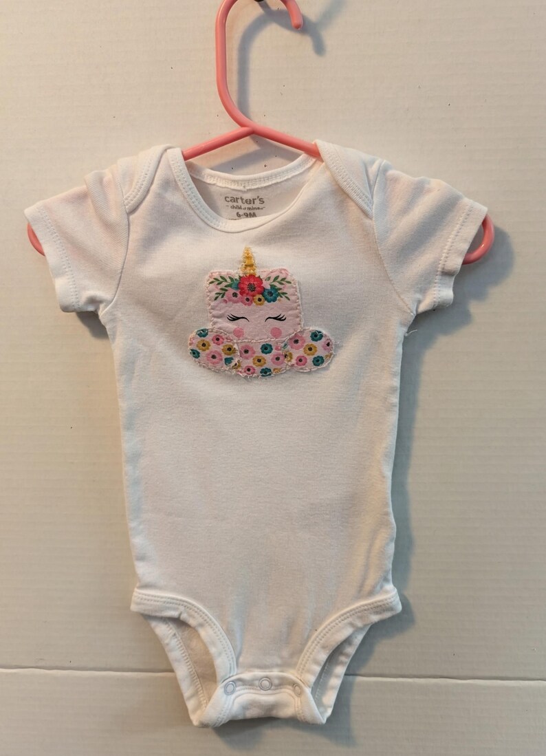 Pode incluir: Body de beb&ecirc; branco com um design de unic&oacute;rnio. O unic&oacute;rnio tem um rosto rosa, um chifre dourado e uma crina floral. O body est&aacute; em um cabide rosa. A marca "Carter's" &eacute; vis&iacute;vel na etiqueta.