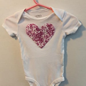 Pode incluir: Body de beb&eacute; branco com um design floral em forma de cora&ccedil;&atilde;o em tons de rosa e roxo. A pe&ccedil;a est&aacute; pendurada num cabide rosa. O nome da marca "Carter's" &eacute; vis&iacute;vel na gola.