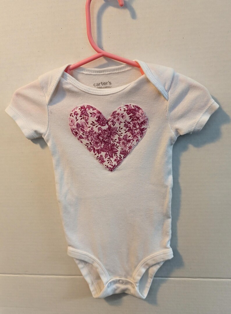 Pode incluir: Body de beb&eacute; branco com um design floral em forma de cora&ccedil;&atilde;o em tons de rosa e roxo. A pe&ccedil;a est&aacute; pendurada num cabide rosa. O nome da marca "Carter's" &eacute; vis&iacute;vel na gola.