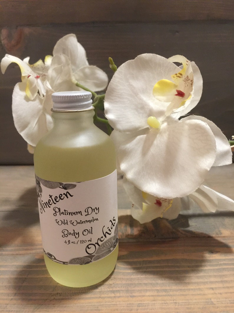 May include: A clear glass bottle with a white label that reads "Nineteen Orchids Platinum Dry Wild Watermelon Body Oil 4 fl oz / 120 ml". The bottle is sitting on a wooden surface with a white orchid flower behind it.
