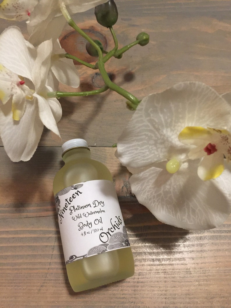 May include: A clear glass bottle with a white label that reads "Nineteen Platinum Dry Wild Watermelon Body Oil 4 fl oz / 120 ml Orchids". The bottle is sitting on a wooden surface with white flowers.