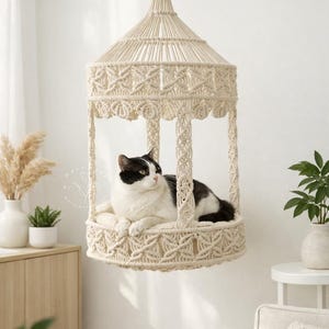 Pode incluir: Uma cama para gatos em macramê creme suspensa no teto. A cama tem uma base circular e um topo em forma de cúpula, com intrincados padrões tecidos. Um gato preto e branco está descansando dentro. Um espaço elegante e confortável para um animal de estimação.