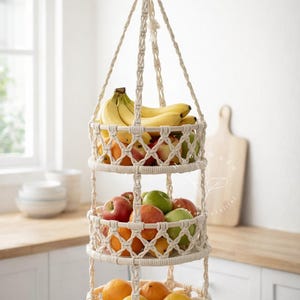 Könnte beinhalten: Ein dreistufiger Hänge-Obstkorb aus cremefarbenem Makramee. Der obere Korb enthält Bananen, der mittlere Korb rote und grüne Äpfel, und der untere Korb ist mit Orangen und Zitronen gefüllt. Der Korb hängt an einem einzigen Seil.