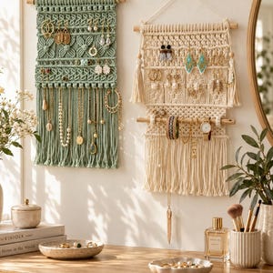 May include: Two wall-mounted macrame jewelry organizers, one sage green and one cream, featuring wooden dowels, intricate knotted patterns, and hanging fringe. These organizers display various earrings, necklaces, bracelets, and a wristwatch.