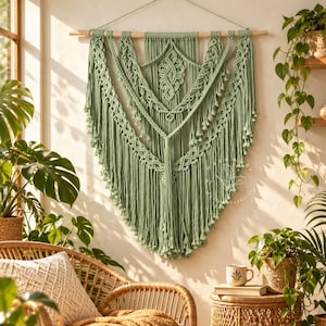 May include: A sage green macramé wall hanging suspended from a horizontal wooden dowel. The piece features intricate knotted patterns, including central diamond shapes and layered fringe, with a subtle watermark logo visible on the right side.