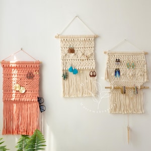 May include: Three macrame jewelry organizers in peach and cream colors hang on a white wall. The organizers have wooden dowels and fringe. A round, woven mirror is also on the wall. Earrings and other jewelry are displayed.