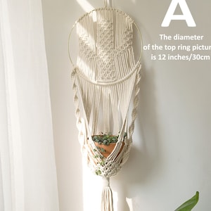 May include: Cream macrame plant hanger with a gold ring, holding a terracotta pot with greenery. The top ring diameter is 30cm. The design includes a woven pattern and a tassel at the bottom, hanging against a white wall.