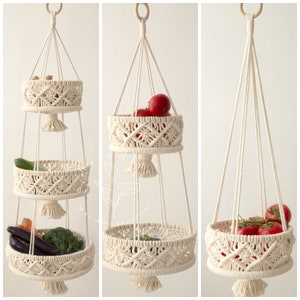 Handcrafted three-tiered macrame fruit basket with natural cotton rope, featuring intricate knotting patterns, tassels, and wooden ring for hanging. filled with apples, eggplants, citrus, and hanging wooden ring
