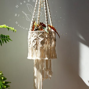 May include: Handmade macrame plant hanger in natural off-white. The hanging planter features a geometric design with a fringed bottom. The plant holder is suspended from a single cord and is filled with a plant with red and green leaves.