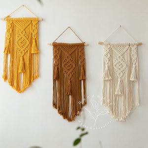 May include: Three macrame wall hangings in mustard yellow, brown, and cream. Each features intricate knotting, wooden dowels, and tassels. The handmade wall art is displayed against a white wall, showcasing the craft.
