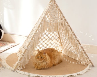 May include: A beige macrame cat teepee with a fringe border and a ginger cat resting inside. The teepee is on a round jute rug with a fringe border. The text "hand made macrame beautiful" is visible in the image.