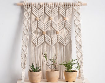 May include: A macrame wall hanging with a wooden shelf, holding three potted plants. The off-white macrame features geometric patterns and wooden accents. The shelf is made of light-colored wood.