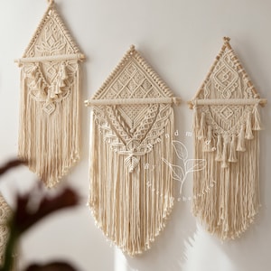 Pode incluir: Três tapeçarias de macramé em forma triangular com uma barra de madeira na parte superior. As tapeçarias são feitas com cordão de algodão de cor natural e têm uma franja na parte inferior. As tapeçarias estão dispostas em uma fila em uma parede branca.