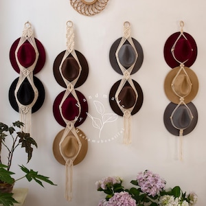 Puede incluir: Organizadores de sombreros montados en la pared con sombreros fedora en varios colores, incluyendo burdeos, negro, marrón y gris. Los sombreros están suspendidos por colgadores de macramé color crema, creando una exhibición elegante y funcional. Una planta y flores están en primer plano.