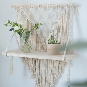 May include: A macrame wall hanging with a wooden shelf. The hanging is made of natural cotton rope and has a diamond pattern. The shelf is made of light wood and is decorated with a vase of white flowers and a potted succulent.