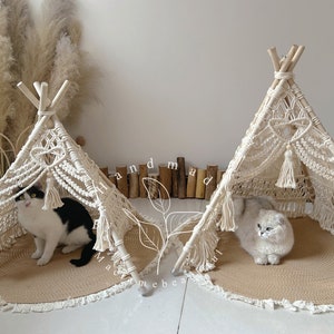 Op de afbeelding: Twee witte macramé katten tipi's met franjes en kwastjes. In elke tipi zit een kat. De tipi's staan op een beige geweven mat met franjes.