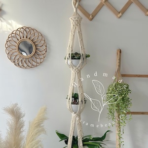 May include: A cream-colored macrame plant hanger with three tiers, each holding a white or light green ceramic pot with greenery. The hanger is suspended from a loop at the top. The background includes a round woven mirror and decorative plants.