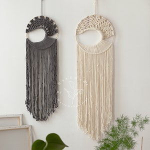 May include: Two macrame wall hangings with a tree of life design. The left one is gray and the right one is cream.