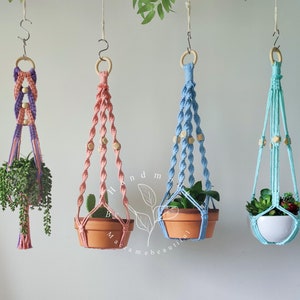 Op de afbeelding: Vier macramé plantenhangers met houten kralen, elk met een potplant. De hangers zijn roze, blauw en paars.