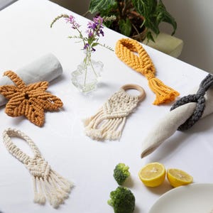 Puede incluir: Una colección de servilleteros de macramé hechos a mano en varios diseños y colores, incluyendo una forma de hoja, un anillo con flecos y un estilo anudado. Los colores incluyen amarillo mostaza, crema y gris. Los artículos están dispuestos sobre un mantel blanco.