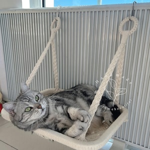 Op de afbeelding: Een grijze tabbykat die ontspant in een witte hangmat van touw. De hangmat hangt aan twee haken aan een witte muur. De tekst "Handmade with love & care, beautiful" is zichtbaar in de afbeelding.