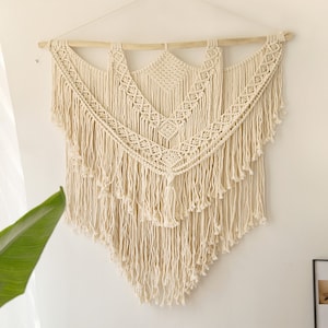 May include: A large, off-white macrame wall hanging with a geometric pattern and fringe. The hanging is made of natural cotton cord and is attached to a wooden dowel.