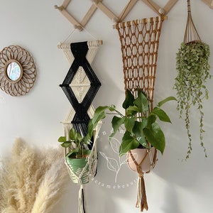 May include: Two macrame plant hangers with a geometric design. The hangers are made with natural fibers and have a rustic look. The hangers are hanging on a white wall with a wooden rack above them. The hangers are holding potted plants.