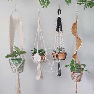 May include: Four macrame plant hangers with different designs and colors. The hangers are made with natural cotton cord and feature a variety of knots and patterns. Each hanger is holding a potted plant.