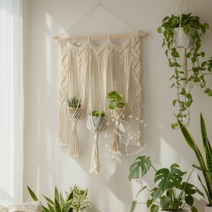 Puede incluir: Un tapiz de pared de macramé color crema con tres jardineras colgantes, cada una con una planta pequeña. El diseño de macramé presenta intrincados nudos y flecos. La habitación también incluye un sofá con cojines decorativos y varias plantas en maceta.