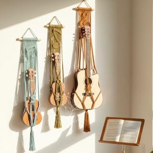 May include: Three stringed instruments, including a guitar and two ukuleles, hang on a white wall in macrame hangers. A wooden music stand holds sheet music. A small potted plant and a stack of books sit on the floor. The room is lit by sunlight.