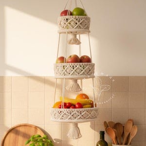 Handmade three-tier macrame fruit basket hanging in a bright kitchen, woven cotton shelves holding apples, bananas, oranges, and tomatoes, boho farmhouse decor for organized countertop storage and natural kitchen styling