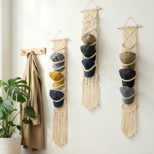 May include: Three macrame hat holders in a neutral tone are suspended on a white wall, each displaying several baseball caps in different colours. A wooden bench and coat rack are also present, offering a stylish storage solution.