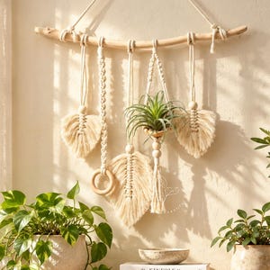 May include: A wall hanging featuring a light-colored wooden branch with cream-colored macrame cords, wooden beads, and fringe leaf shapes. A small air plant is suspended in a macrame holder, and a wooden ring is attached to one of the cords.