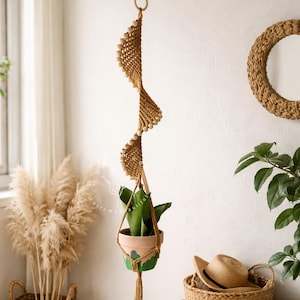 Puede incluir: Un colgador de plantas de macramé hecho a mano con un diseño en espiral, que sostiene una planta en maceta. El macramé beige está adornado con cuentas de madera. La imagen también presenta una cesta tejida, un banco de madera y un espejo decorativo, creando una estética bohemia.