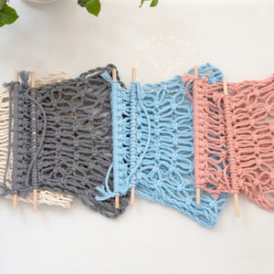 Puede incluir: Cinco tapices de macram&eacute; en crema, gris, azul y rosa, cada uno con un dise&ntilde;o de tejido abierto &uacute;nico. Las piezas de macram&eacute; est&aacute;n unidas a clavijas de madera. La decoraci&oacute;n de pared hecha a mano se exhibe sobre un fondo blanco.