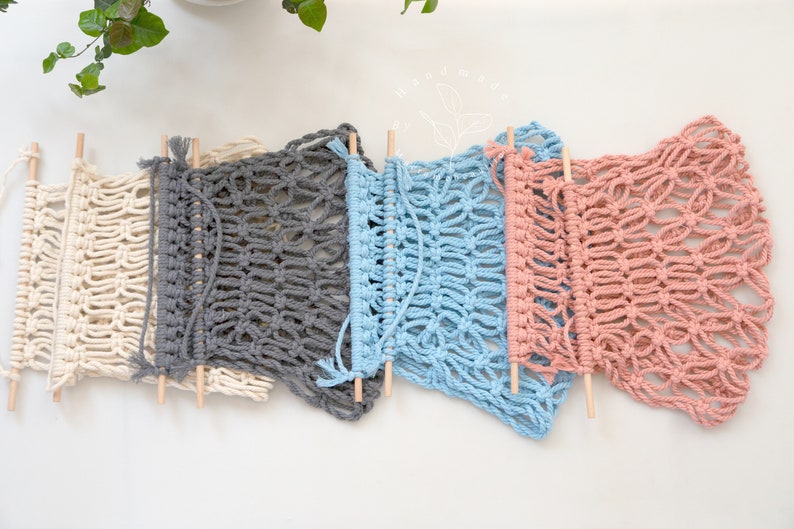 Puede incluir: Cinco tapices de macram&eacute; en crema, gris, azul y rosa, cada uno con un dise&ntilde;o de tejido abierto &uacute;nico. Las piezas de macram&eacute; est&aacute;n unidas a clavijas de madera. La decoraci&oacute;n de pared hecha a mano se exhibe sobre un fondo blanco.
