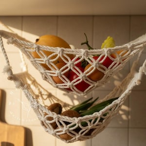 Puede incluir: Hamaca de frutas de macram&eacute; de dos niveles, color crema, colgando del techo. La hamaca superior contiene mangos, tomates y un pimiento amarillo. La hamaca inferior contiene aguacates y jud&iacute;as verdes. Escena de cocina con una tabla de cortar de madera.