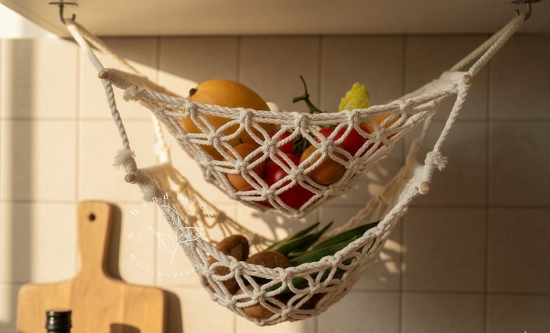 Puede incluir: Hamaca de frutas de macram&eacute; de dos niveles, color crema, colgando del techo. La hamaca superior contiene mangos, tomates y un pimiento amarillo. La hamaca inferior contiene aguacates y jud&iacute;as verdes. Escena de cocina con una tabla de cortar de madera.