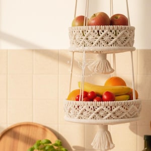 Handmade three-tier macrame fruit basket hanging in a bright kitchen, woven cotton shelves holding apples, bananas, oranges, and tomatoes, boho farmhouse decor for organized countertop storage and natural kitchen styling