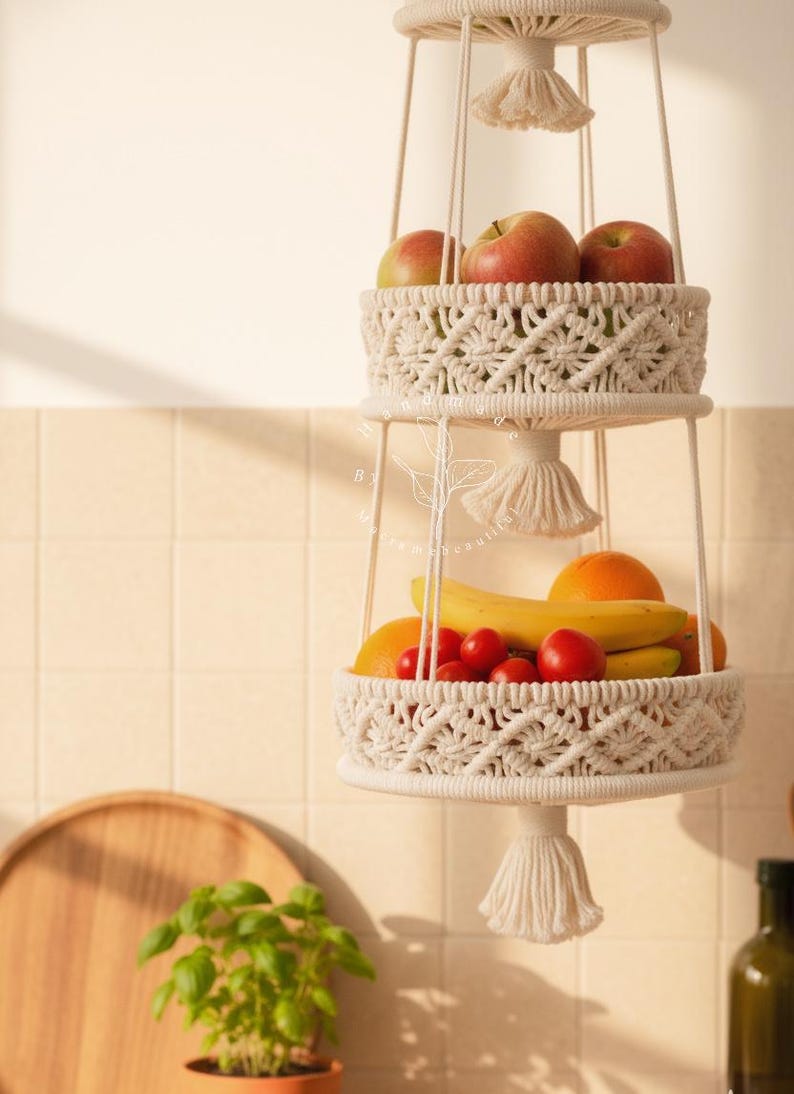 Handmade three-tier macrame fruit basket hanging in a bright kitchen, woven cotton shelves holding apples, bananas, oranges, and tomatoes, boho farmhouse decor for organized countertop storage and natural kitchen styling