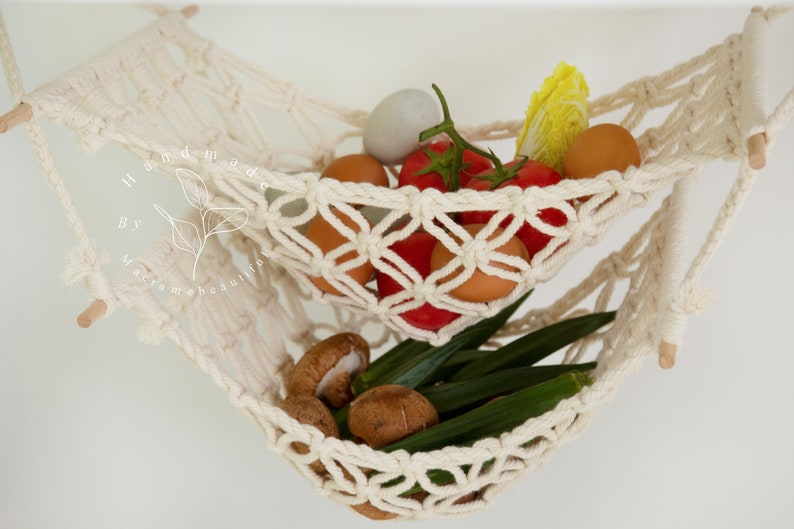 Overhead view of a two-tiered macrame fruit hammock filled with fresh bananas, tomatoes, and apples, showcasing the detailed knotwork, providing a stylish and bohemian kitchen storage solution