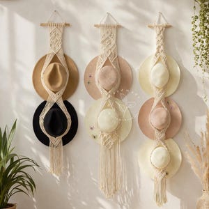 May include: Three macrame hat holders with hats hanging on a white wall. The hats are in various colors, including black, tan, and pink. The macrame is cream-colored, and the holders are attached to wooden dowels.