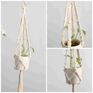 May include: Cream-colored macrame plant hanger with wooden beads and a tassel. The plant hanger suspends a small white ceramic pot with a green plant inside. The hanger is suspended from a wooden ring.