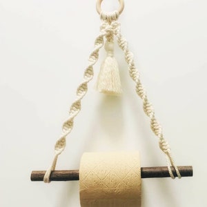 K&ouml;nnte beinhalten: Ein wei&szlig;er Makramee-Toilettenpapierhalter mit einem Holzstift und einem Quasten. Der Halter h&auml;ngt an einem Holzring.