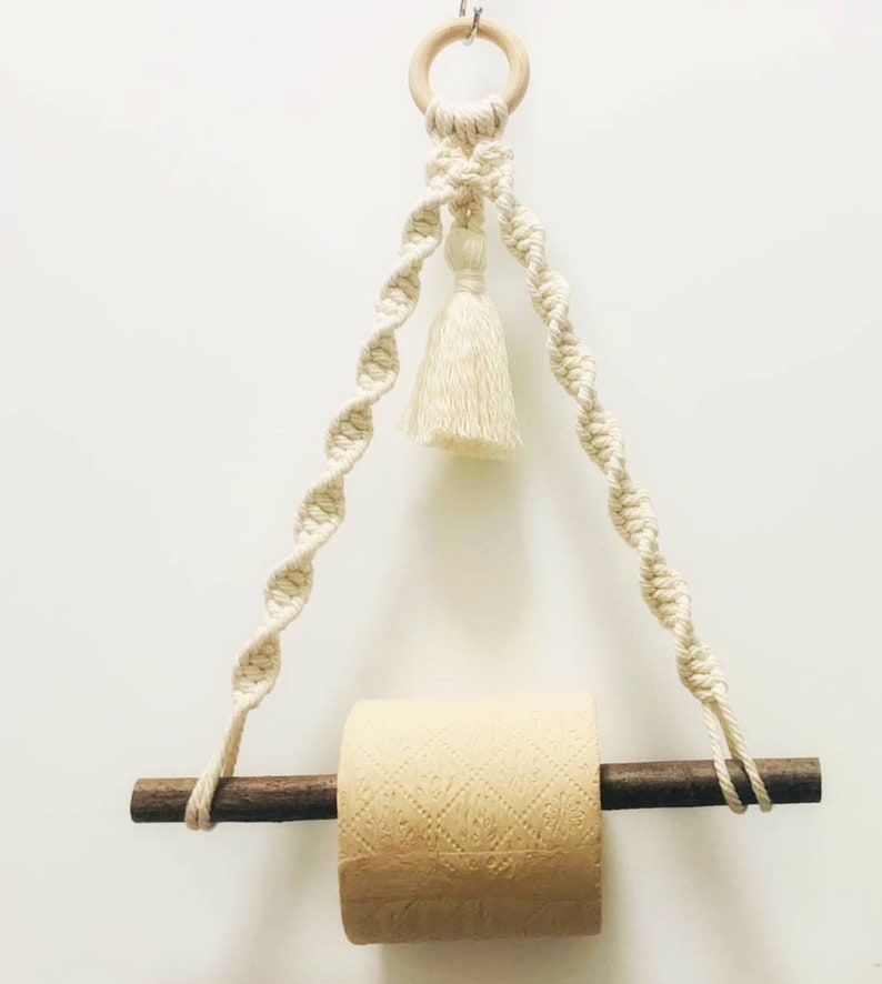 K&ouml;nnte beinhalten: Ein wei&szlig;er Makramee-Toilettenpapierhalter mit einem Holzstift und einem Quasten. Der Halter h&auml;ngt an einem Holzring.
