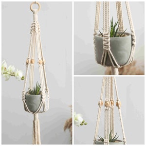 May include: A macrame plant hanger with a small, gray pot holding a succulent plant. The off-white rope is knotted and features wooden beads. A wooden ring is at the top for hanging. The plant hanger is a decorative item for indoor plants.