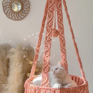 May include: A peach-colored macrame cat bed with a white cat inside. The hanging bed is suspended from a hook and features intricate knotting and a fringed edge. The words "handmade by macramebeautiful" are visible.