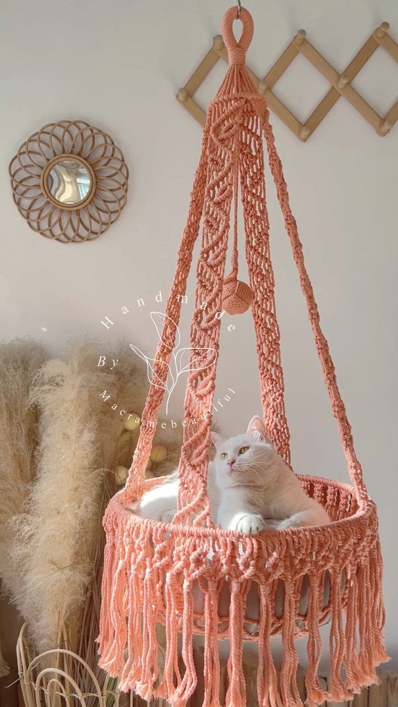 May include: A peach-colored macrame cat bed with a white cat inside. The hanging bed is suspended from a hook and features intricate knotting and a fringed edge. The words "handmade by macramebeautiful" are visible.