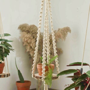 May include: A white macrame plant hanger with a wooden round shelf and a green plant in a terracotta pot. The hanger is 43.3 inches/110cm long, including the tassels.