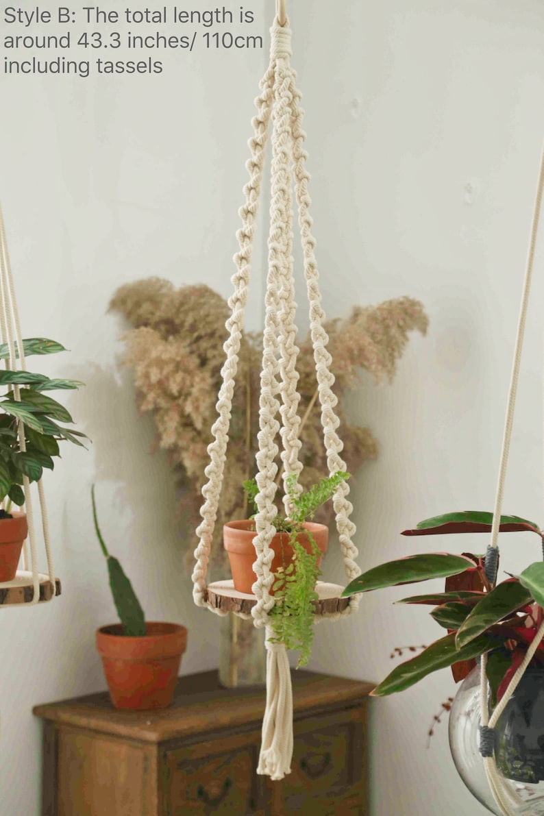 May include: A white macrame plant hanger with a wooden round shelf and a green plant in a terracotta pot. The hanger is 43.3 inches/110cm long, including the tassels.